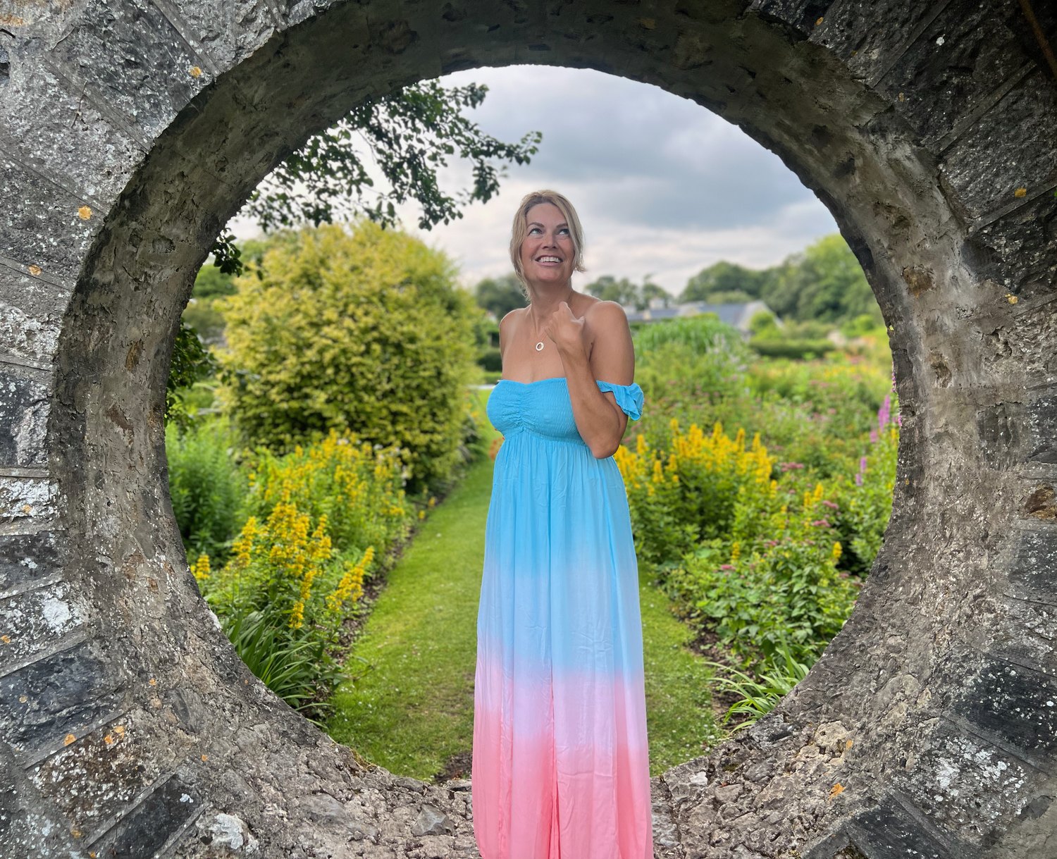 Michelle Tucker standing in a circular stone garden archway, with flower borders and a lawn beyond
